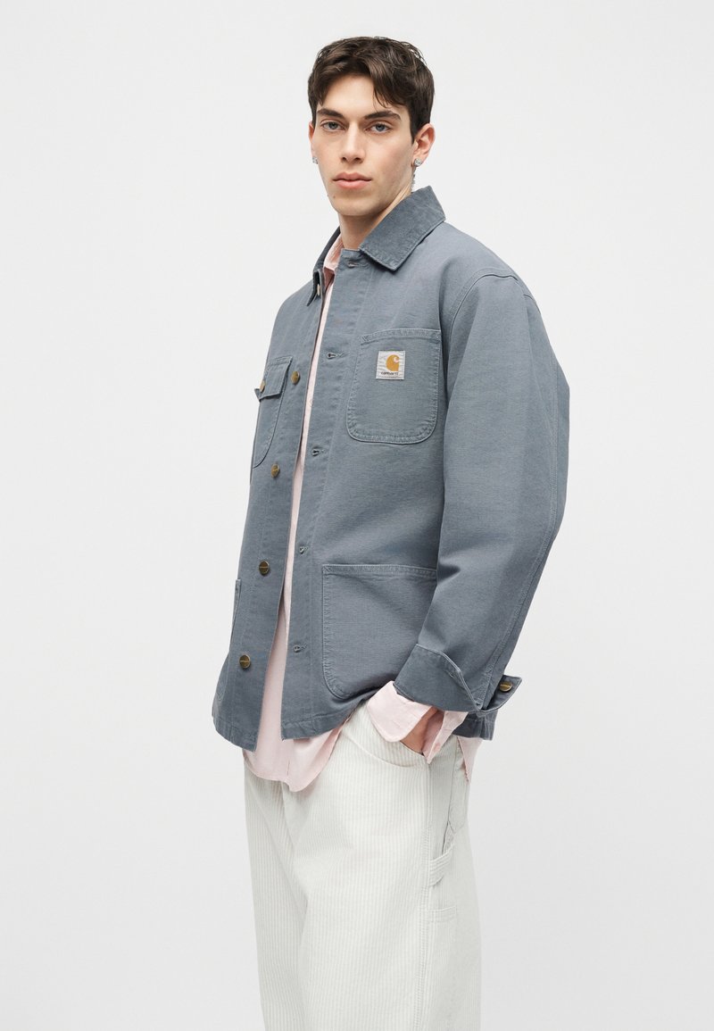 Giovane uomo che indossa una giacca da lavoro blu con bottoni e tasche, una camicia rosa chiaro e pantaloni bianchi, in piedi con le mani nelle tasche.