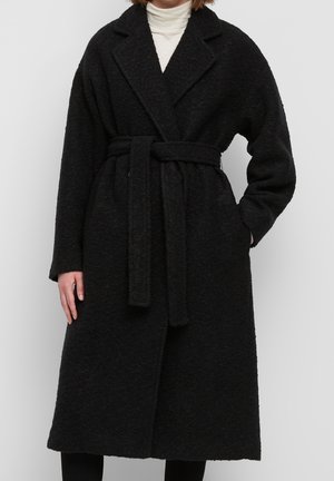 Frau trägt einen langen schwarzen texturierten Mantel mit Gürtel über einem weißen Rollkragenpullover, Hände in den Taschen, steht vor einem einfarbigen Hintergrund.