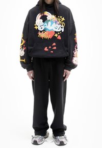 Sudadera negra con gráficos coloridos, incluyendo corazones y nubes, combinada con pantalones de chándal negros a juego y zapatillas multicolores.