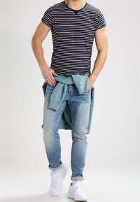 Marinblå och vitrandig bomullströja, ljusblå denimjeans och en ljus tealtröja knuten runt midjan. Vita sneakers synliga.