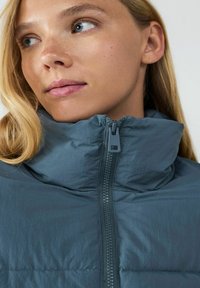 Szara kurtka ocieplana z wysokim kołnierzem, pełnym zamkiem błyskawicznym i teksturowanym materiałem. Zauważalny detal zamka z gładkim wykończeniem.