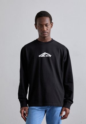 Schwarzes Langarmshirt aus Baumwolle mit weißem Grafikdruck. Mit Rundhalsausschnitt und lässigem Schnitt, kombiniert mit hellen Denim-Jeans.