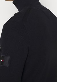 Granatowy sweter z golfem o prążkowanej teksturze, dopasowanym kroju i naszywce z logo na rękawie. Posiada detale paneli na ramionach.