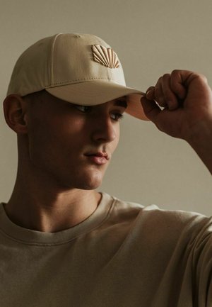 Beige baseball cap with a shell emblem, made of soft fabric, adjustable strap, worn slightly tilted, paired with a light-colored shirt.