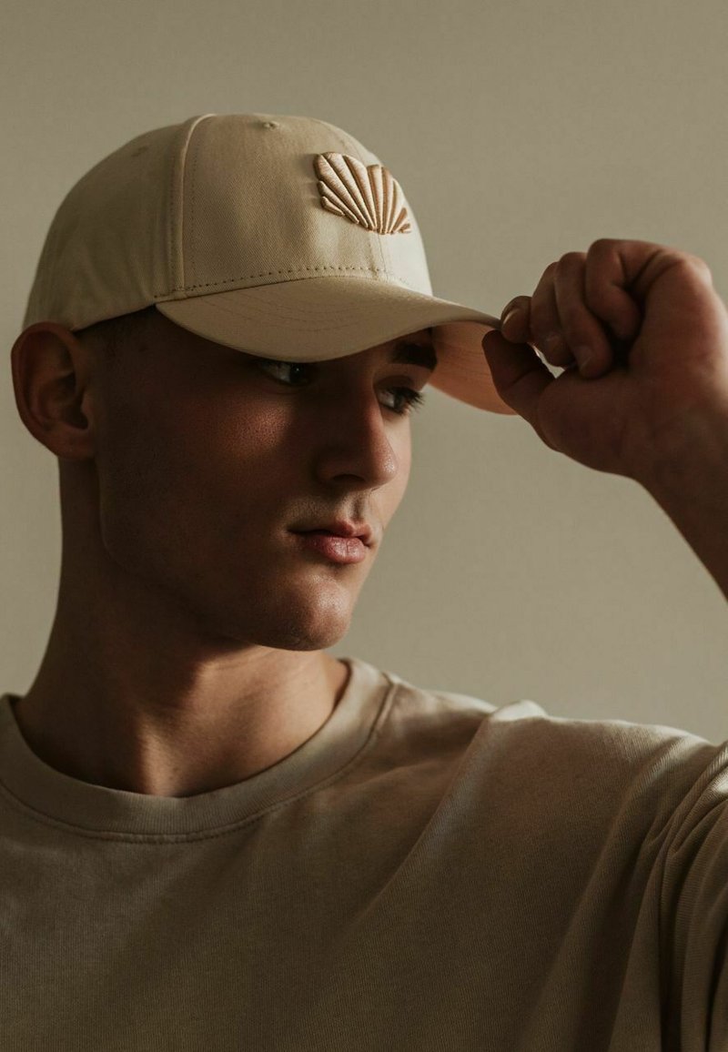 Beige színű baseball sapka kagyló emblémával, puha anyagból, állítható pánttal, enyhén ferde módon viselve, világos színű inggel párosítva.