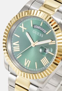 Ceas Guess cu un cadran verde, bratara din aur si argint, afisaj de data si marcaje cu numere romane. Dispune de o bezel texturat din aur.