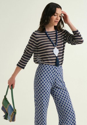 Woman wearing navy striped top and blue patterned pants, holding a blue and green handbag, with a blue beaded necklace and pendant.