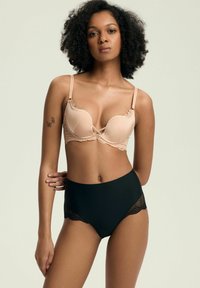 Frau mit lockigem Haar, die einen nude-farbenen Spitzen-BH und schwarze Unterwäsche mit hoher Taille und Spitzenverzierungen trägt, steht vor einem einfarbigen hellen Hintergrund.