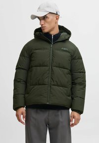 Olivgrüne Pufferjacke mit Kapuze, Reißverschluss, gestepptem Design, elastischen Bündchen und Branding auf der Brust. Über einem schwarzen Oberteil getragen.