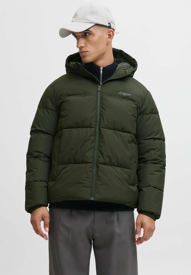 Olivgrüne Pufferjacke mit Kapuze, Reißverschluss, gestepptem Design, elastischen Bündchen und Branding auf der Brust. Über einem schwarzen Oberteil getragen.