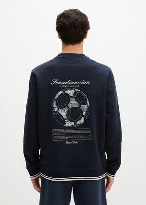 Mann kledd i marineblå genser med slitt fotballgrafikk og teksten "Scandinavian Football Community" på baksiden, stående foran hvit bakgrunn.