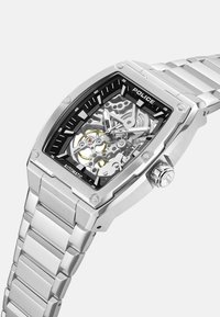 Kwadratowy zegarek ze stali nierdzewnej z przezroczystym szkieletowym cyferblatem, czarnymi akcentami i srebrną bransoletą ogniwową. Posiada wypolerowane powierzchnie i solidne elementy.