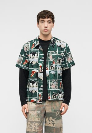 Jeune homme portant une chemise verte à manches courtes imprimée de bandes dessinées par-dessus une chemise noire à manches longues et un pantalon cargo patchwork, debout devant un fond blanc.