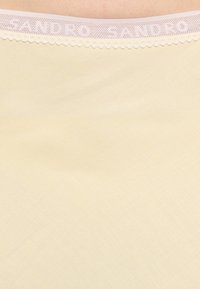 Gros plan sur un tissu de sous-vêtements beige avec une ceinture élastique blanche présentant un texte subtil "SANDRO" et une bordure festonnée.