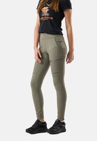 Kvinde iført olivengrønne, tætsiddende cargobukser, sort grafisk t-shirt og sorte sneakers, stående mod en hvid baggrund.