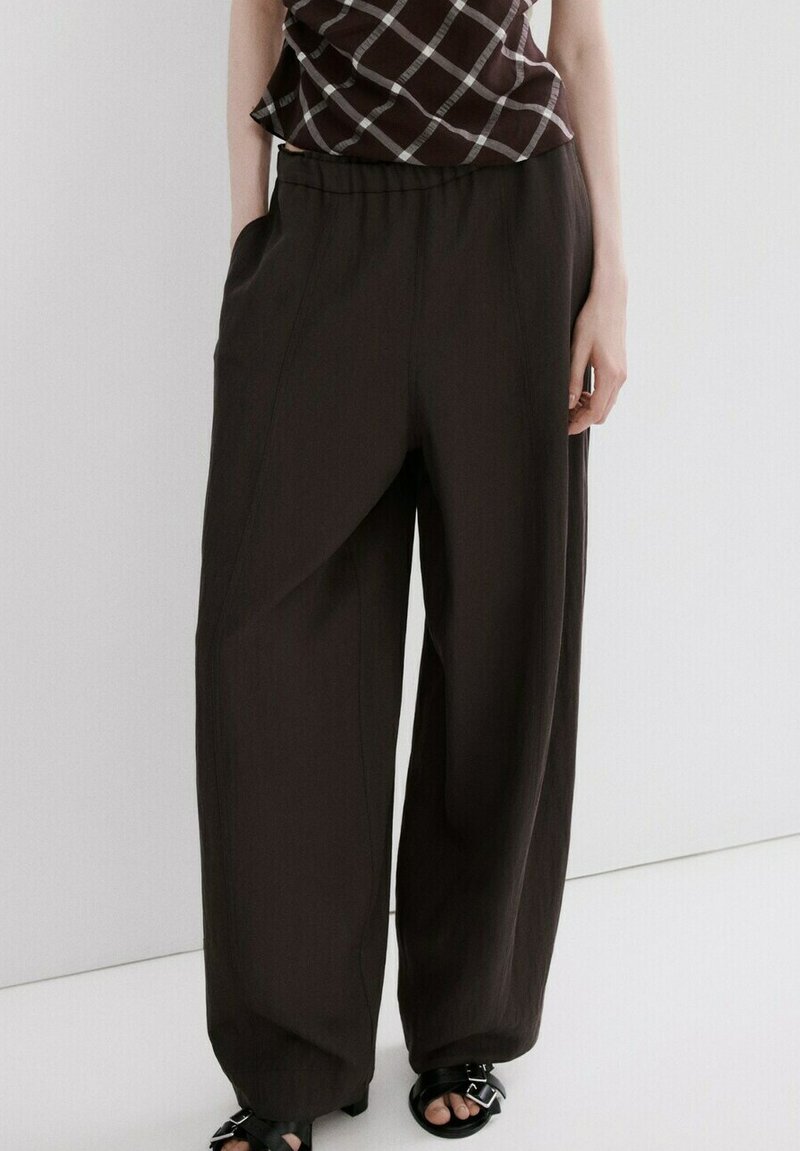 Personne portant un pantalon ample marron foncé, des sandales noires à bouts ouverts et un haut sans manches à motif à carreaux marron et blanc.