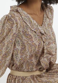 Blus med blommönster i ljusbeige, med en volangkrage, puffärmar och en figurnära midja med ett cremfärgat bälte.