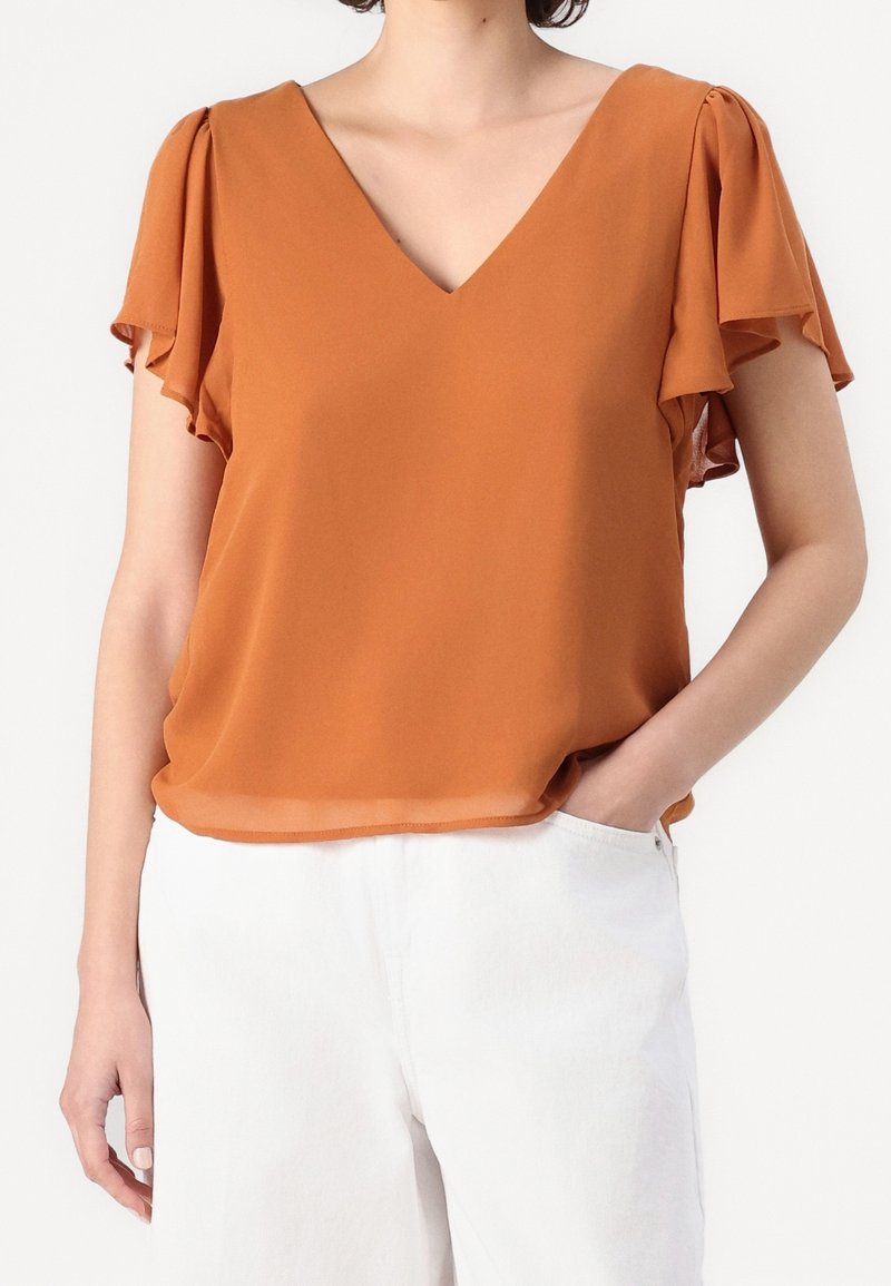 Personne portant un chemisier orange rouille à col en V avec des manches volantes et un pantalon blanc, une main dans la poche.