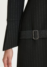 Schwarzes Kleidungsstück mit silbernen Nadelstreifen, versehen mit einem breiten Gürtel mit einer rechteckigen Schnalle, die mit kleinen Nieten und einem strukturierten Stoff geschmückt ist.