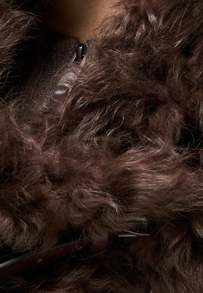 Primo piano di una giacca di pelliccia riccia marrone scuro con cerniera metallica visibile e parte di una fascia in pelle su un sottogiacca di maglia scura.