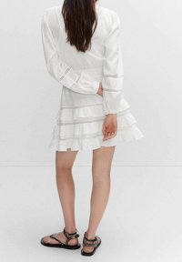 Robe blanche à étages avec manches longues, présentant des accents de coutures horizontales. Portée avec des sandales plates noires ornées de petits clous.