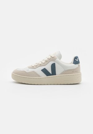 White leather sneakers with grey suede accents and a blue logo. Features a textured toe and a cushioned sole. Minimalist design.