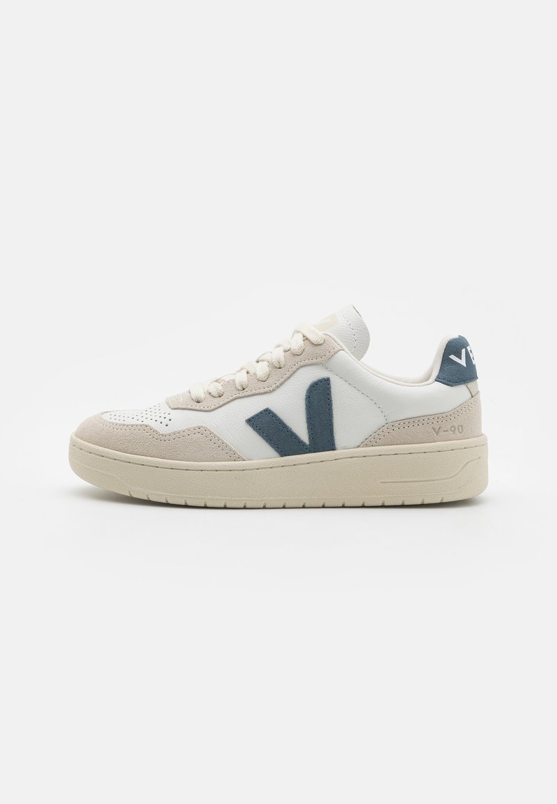 Sneaker in pelle bianca con dettagli in suede grigio e logo blu. Presenta una punta testurizzata e una suola imbottita. Design minimalista.