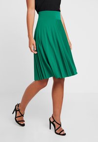 Gonna plissettata verde con una fascia larga in vita, lunghezza al ginocchio, design fluido, realizzata in un tessuto morbido, abbinata a tacchi a strappo neri.