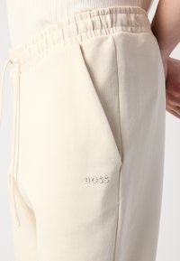 Close-up van beige joggers met elastische tailleband, zijzak, trekkoord en klein "BOSS" logo nabij de zak.