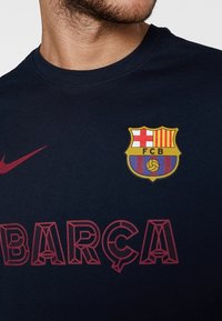 Navy blue cotton T-shirt with round neckline, featuring a yellow and red FC Barcelona crest and red “BARÇA” text with dotted pattern.