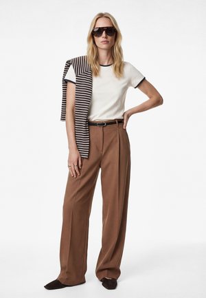 Frau trägt übergroße braune Hose, weißes T-Shirt mit marineblauem Rand, gestreiften Pullover über der Schulter, dunkle Sonnenbrille und schwarze Schuhe, steht mit einer Hand in der Hüfte.