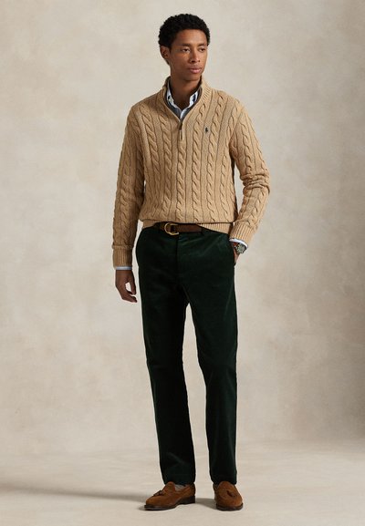 Jonge man draagt een beige kabelgebreide trui, donkergroene broek, bruine riem en bruine schoenen met kwastjes, staand tegen een egale achtergrond.