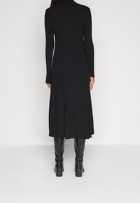 Vestito lungo a maniche lunghe in maglia nera a coste, con collo alto, orlo svasato e silhouette aderente, abbinato a stivali neri sopra il ginocchio.