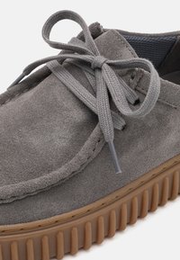 Gray suede shoe with round toe, contrasting brown rubber sole, and flat laces. Features textured surface and visible stitching accents.