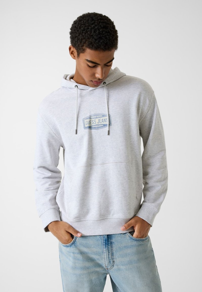 Szara bluza z kapturem wykonana z miękkiego materiału, posiadająca kieszeń z przodu, kaptur z regulacją sznurkiem oraz niebieską naszywkę z napisem "GUESS JEANS".