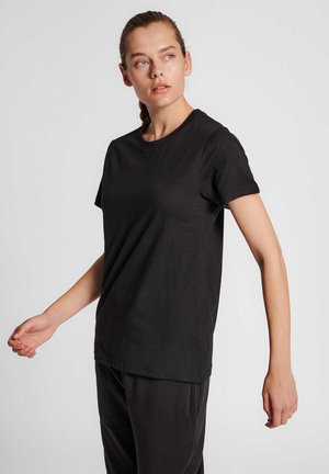 Młoda kobieta ubrana w prostą czarną koszulkę i spodnie, patrzy w lewo z lekko uniesionymi ramionami na jasnym tle.