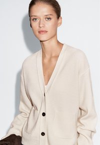 Beige geribde cardigan met een V-hals, voorzakken en bruine knopen. Zachte textuur en relaxte pasvorm, gedragen over een witte top.