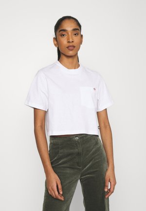 T-shirt bianco corto con maniche corte e taschino sul petto. Indossato con pantaloni in corduroy verde dotati di tasche laterali e un bottone frontale.