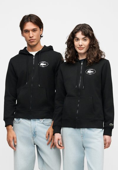 Мъж и жена стоят един до друг, носещи черни суичъри с цип с логота Lacoste Paris и светлосини дънки на едноцветен фон.
