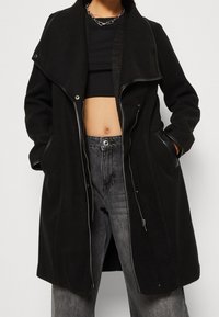 Svart ullblandad kappa med överdimensionerad krage, dragkedja framtill och huva. Har sidofickor och läderliknande detaljer. Texturerad och strukturerad design.