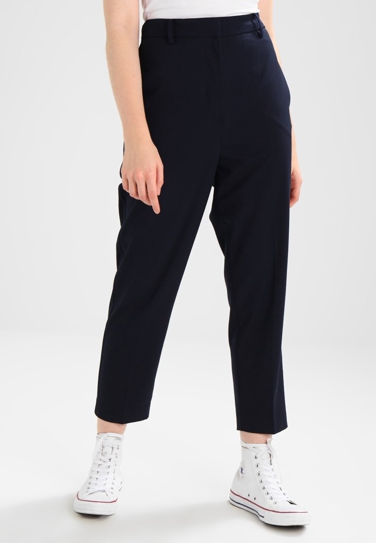 Personne portant un pantalon noir court, des baskets blanches montantes et un haut blanc, debout devant un fond clair uni.