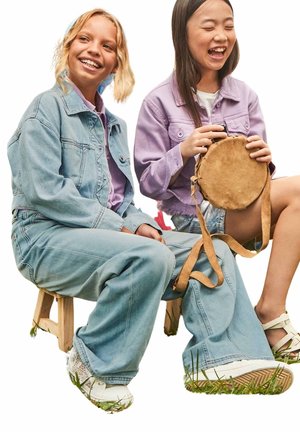 Due bambini seduti che indossano giacche di jeans, jeans azzurri e scarpe bianche. Uno tiene una borsa rotonda color beige con una tracolla. Lo sfondo è bianco.