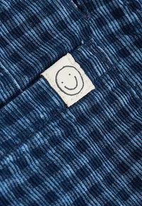 Biała metka z prostą czarną uśmiechniętą buźką naszyta na teksturowanej, ciemnoniebiesko-jasnoniebieskiej tkaninie w paski.