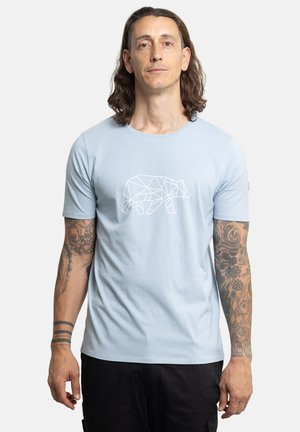Uomo con capelli lunghi e braccia tatuate che indossa una maglietta azzurro chiaro con un disegno geometrico bianco di un orso, in piedi davanti a uno sfondo semplice.