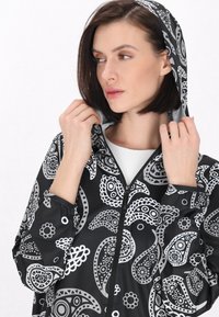 Femeie care își ajustează gluga unei jachete negre cu modele paisley alb și gri, privindu-se lateral pe un fundal simplu de culoare deschisă.