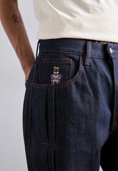 Dark blue denim jeans featuring an embroidered cartoon character on the front pocket. Visible stitching in contrasting orange.