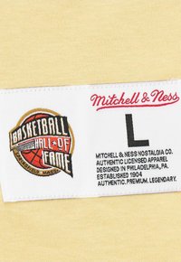 Gelbes Stoffetikett mit eingesticktem "Basketball Hall of Fame" Logo und dem Namen "Mitchell & Ness"; umfasst die Größe "L" und Produktdetails.