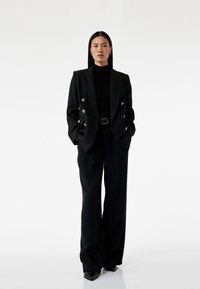 Zwarte dubbelgeknoopte blazer met metalen knopen, gecombineerd met een aansluitende zwarte coltrui en brede zwarte broeken, afgemaakt met puntige hakken.