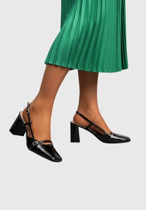 Jambes en collants couleur bronzée portant des escarpins à bride arrière en cuir verni noir avec des talons blocs et des boucles, associés à une jupe midi plissée verte.