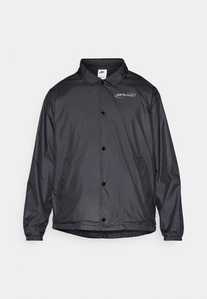 Black windbreaker jacket with a collared neck, snap buttons, elastic cuffs, side pockets, and a logo on the left chest. Lightweight fabric.